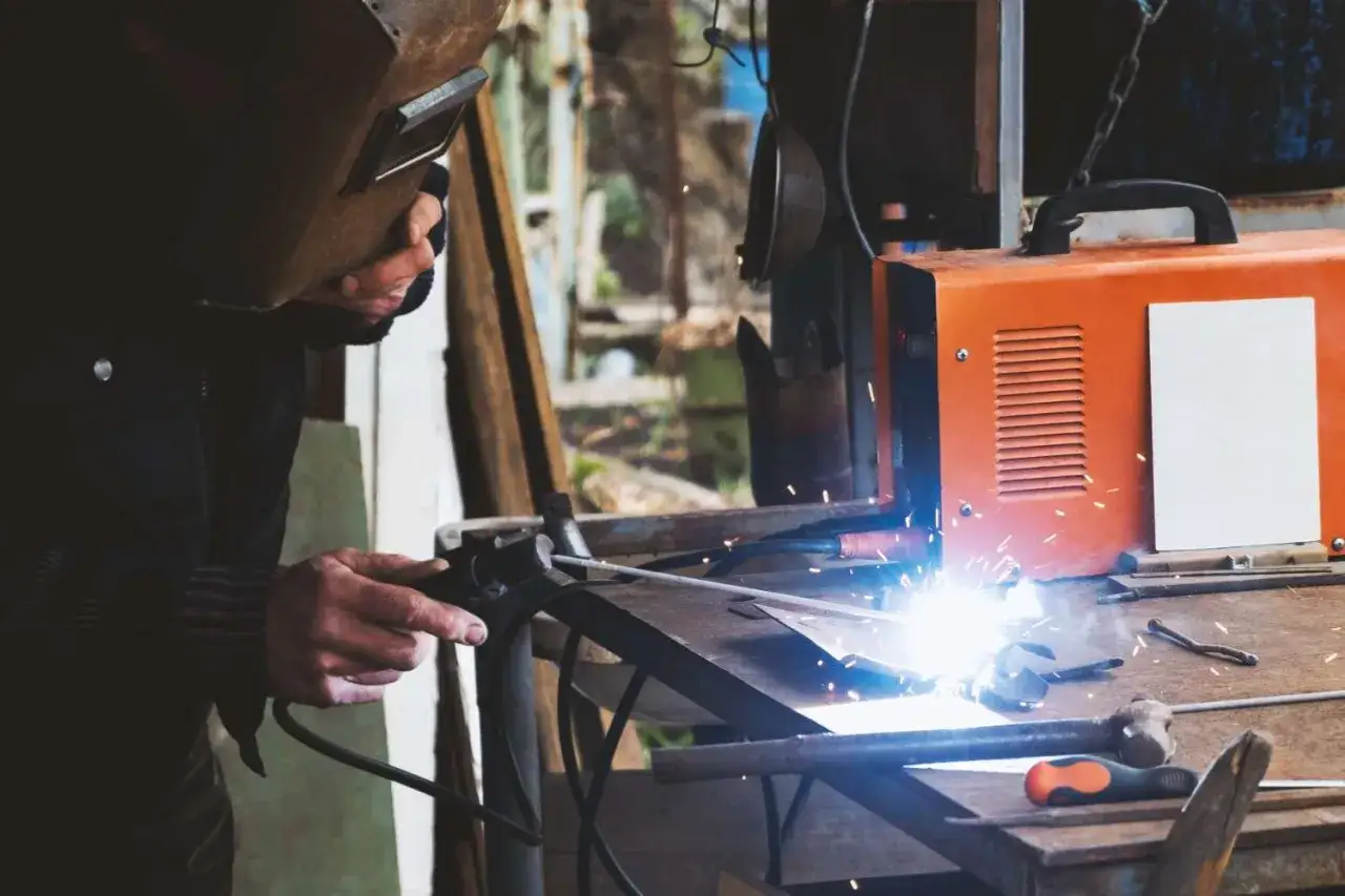 Soldador trabajando con una máquina naranja, creando chispas. Se aprecian varios tipos de soldadores en el taller.