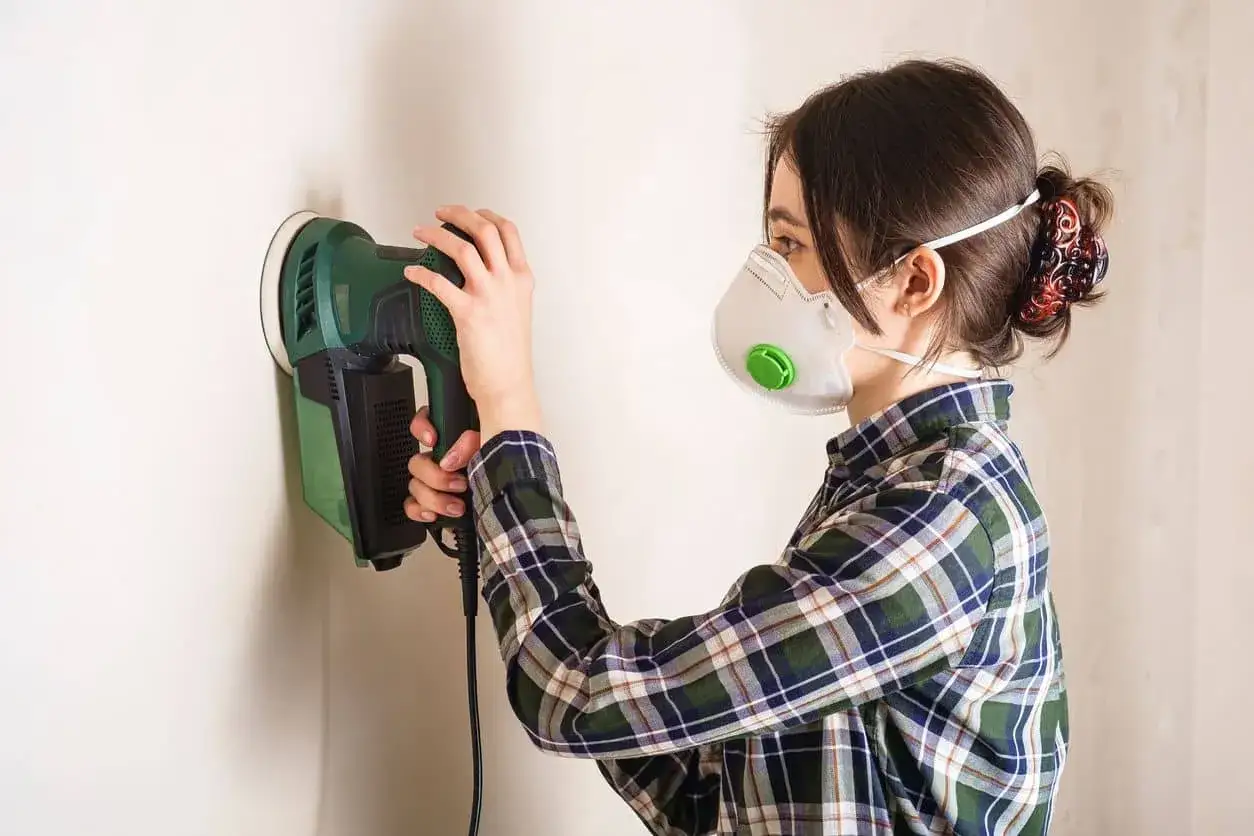 Mujer con mascarilla lijando una pared con una lijadora eléctrica.