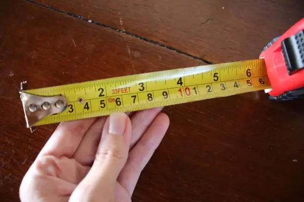 Una mano sujeta una cinta métrica amarilla sobre una mesa de madera. La cinta muestra medidas en pies y metros, demostrando para que sirve la cinta métrica: medir distancias.