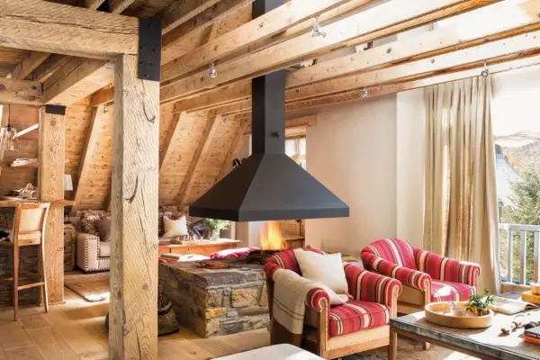 Chimenea central de diseño moderno, uno de los tipos de chimeneas que aporta calidez a este salón rústico con vigas de madera.