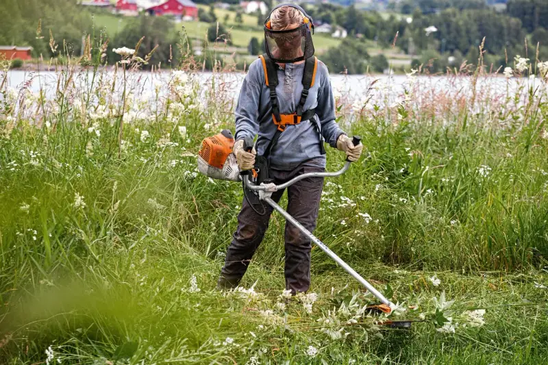 Persona con equipo de protección usa desbrozadora para cortar hierba alta, mostrando la diferencia entre desbrozadora y cortabordes en un paisaje rural.